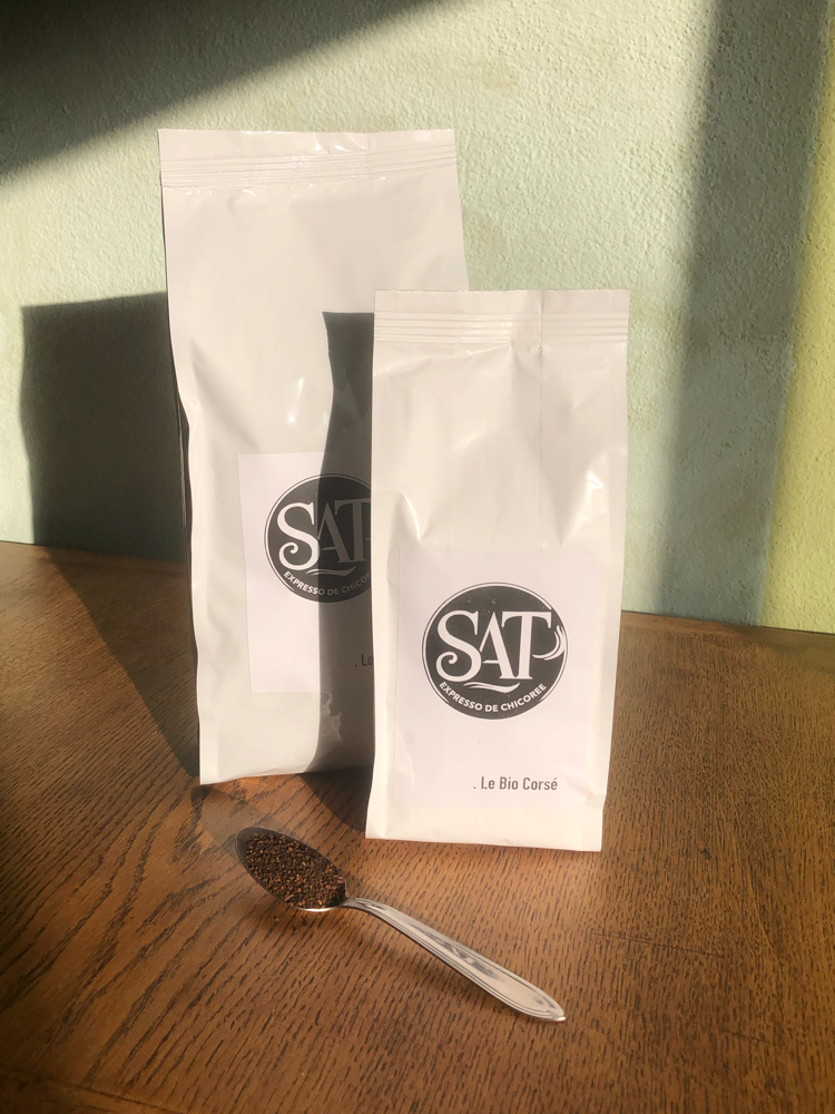 Deux sacs de SAT Bio Corsé Expresso de chicorée sur table en bois, mur vert en arrière-plan, cuillère à café remplie de chicorée posée devant