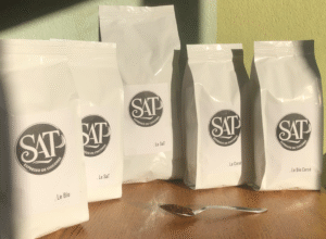 Quatre sacs de 400 g et un sac de 1 kg de SAT Expresso de chicorée posés sur une table en bois éclairée par le soleil devant un mur vert