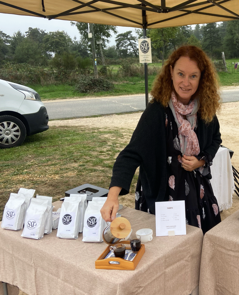 Anne Marie servant du Sat sur le stand du marché en plein air de Chambles, avec des paquets de SAT Chicorée exposés sur une table.